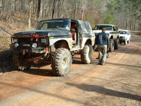 1992 Ford Mustang - A line of off-road vehicles, led by a heavily modified black Ford Bronco, travels down a dirt road.