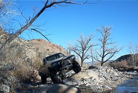 1986 Ford Bronco - Pickup truck navigating a rocky stream bed under a clear blue sky.