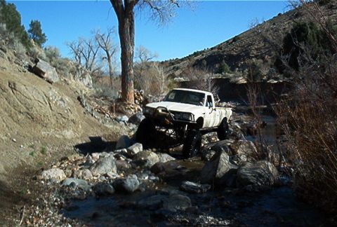 1986 Ford Bronco - A white pickup truck is shown off-roading through a rocky stream bed under a clear blue sky.