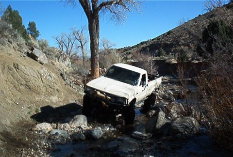 1986 Ford Bronco - Pickup truck off-roading through a rocky stream bed under a clear blue sky.