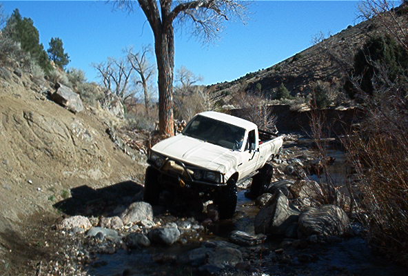 White pickup truck navigating a rocky stream bed with a steep, dry hillside in the background