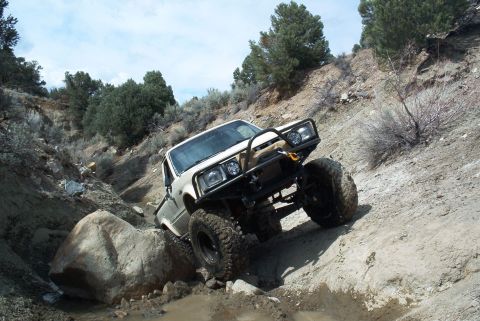 1986 Ford Bronco - Modified white truck navigates a challenging off-road course with large boulders and steep terrain.