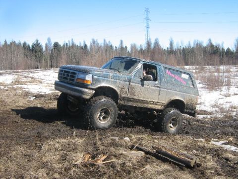 1995 Ford Bronco - A heavily modified Ford Bronco navigates a challenging muddy terrain under a clear blue sky.