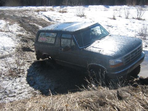 1995 Ford Bronco - This dark SUV appears to be from the early to mid-1990s, possibly a Ford Bronco, struggling through a muddy and snowy terrain.