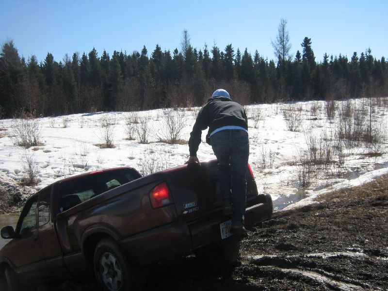Person climbing onto the tailgate of a red pickup truck stuck in mud with snow on the ground and trees in the background