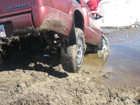 1995 Ford Bronco - A red Chevrolet pickup truck is stuck in a muddy puddle, with mud splattered on its tires and body.
