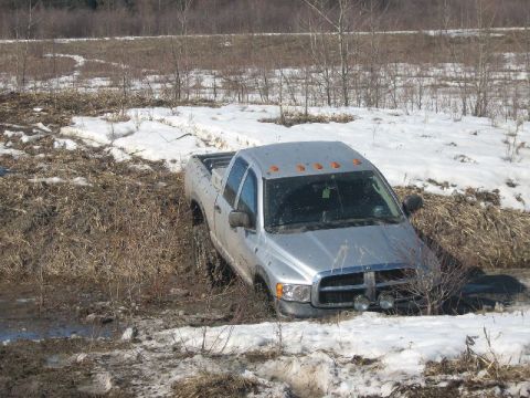 1995 Ford Bronco - A silver pickup truck is stuck in a muddy, snow-covered ditch.