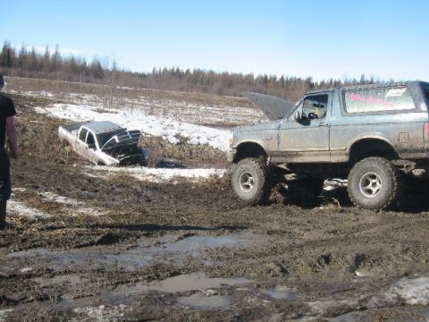 1995 Ford Bronco - Two vehicles are stuck in a muddy field with snow patches and trees in the background.