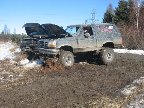 1995 Ford Bronco - Two muddy trucks with hoods open are stuck in a muddy field with snow patches under a clear blue sky.