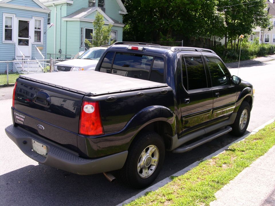 Black pickup truck parked on street, another vehicle behind it. Has a bed cover and roof rack
