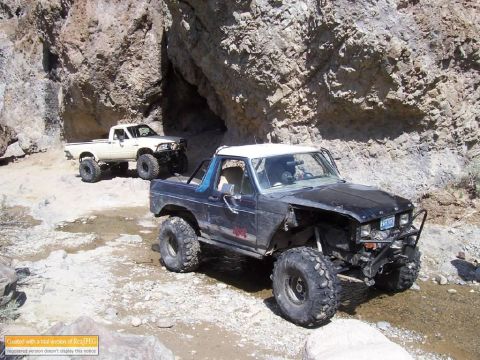 1986 Ford Bronco - Two modified off-road vehicles navigate a rocky, narrow canyon path.