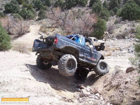 1986 Ford Bronco - Two modified trucks navigate a rocky, off-road trail in a dry, hilly landscape.