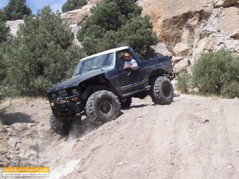 1986 Ford Bronco - Modified Ford Bronco navigates a steep, dusty off-road incline with rocky terrain and sparse vegetation.