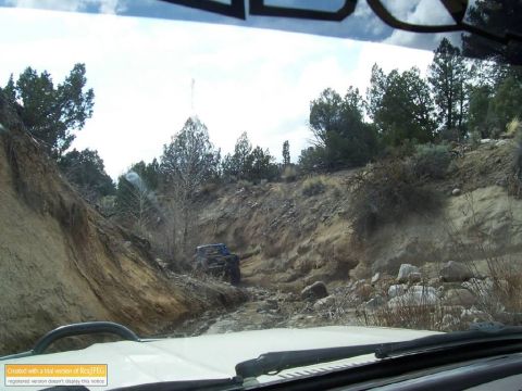 1986 Ford Bronco - Two off-road vehicles navigate a challenging rocky trail through a dry, wooded landscape, as seen from the perspective of another vehicle.