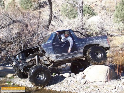 1986 Ford Bronco - A man in a hat navigates a modified SUV through a rocky, off-road terrain with a stream in the foreground.