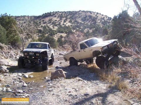 1986 Ford Bronco - Two heavily modified pickup trucks are shown off-roading in a rocky, arid landscape.