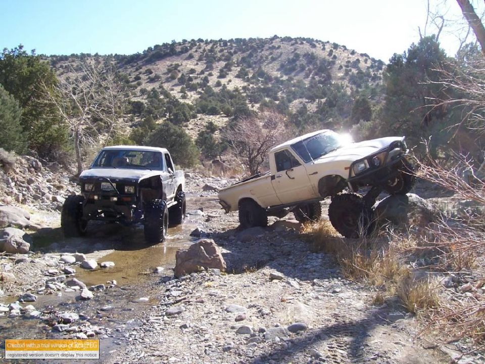 Two lifted pickup trucks navigating a rocky, dry creek bed
