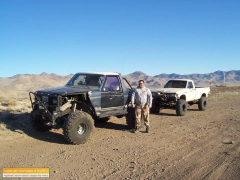 1986 Ford Bronco - A man stands between two modified off-road trucks in a desert setting, with mountains in the background.