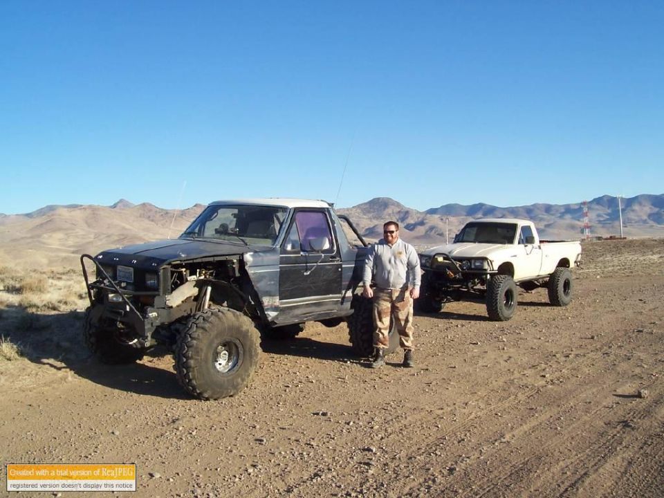 Modified black truck with large tires and a white roof, parked in a desert landscape with another white truck behind it