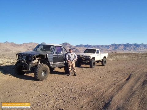 1986 Ford Bronco - Two modified trucks and a man stand in a desert landscape under a clear blue sky.