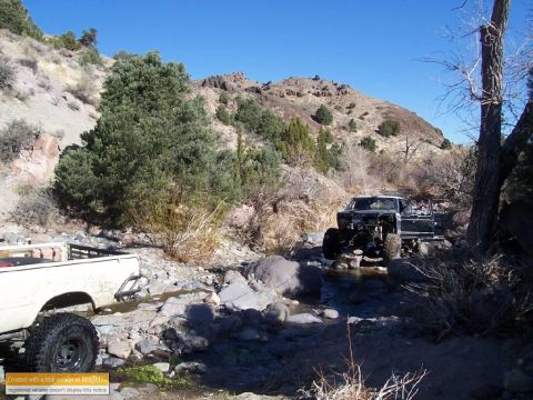 1986 Ford Bronco - Two modified trucks navigate a rocky stream bed under a clear blue sky.
