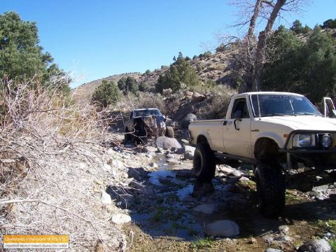 1986 Ford Bronco - Two modified pickup trucks navigate a challenging off-road trail with rocky terrain and water.