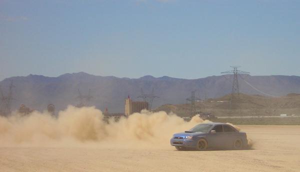 Blue Subaru Impreza sedan throwing up a big dust cloud on a desert flat with mountains behind