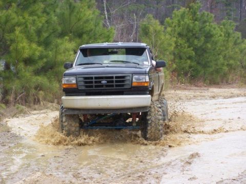 1996 Ford F-150 - This lifted truck is navigating a challenging muddy terrain, kicking up water as it goes.