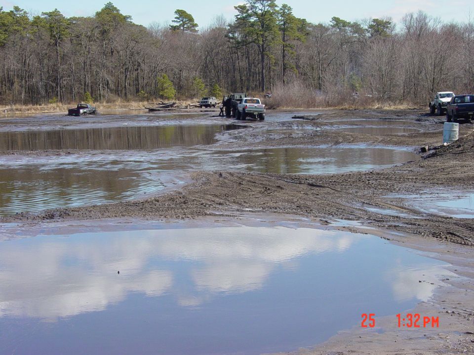 Several trucks are stuck in a muddy area with trees in the background. A water puddle reflects the sky