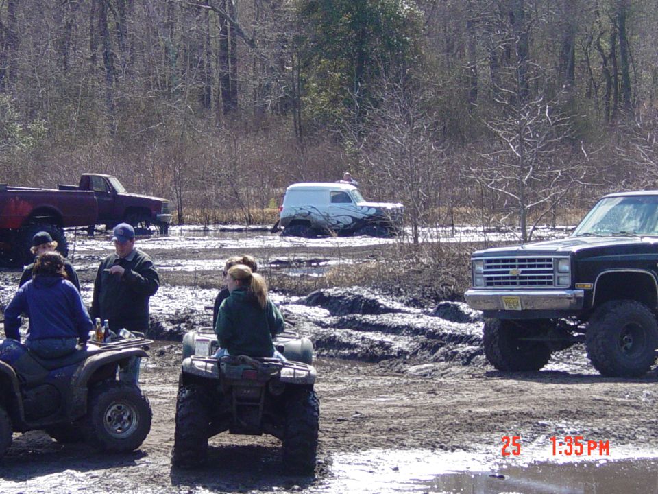 Muddy scene with a lifted black pickup truck in the foreground, people on ATVs, and a maroon truck and white van stuck in mud in the background