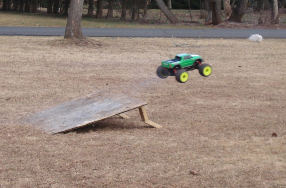 Green remote control monster truck airborne over dry grass near a wooden ramp