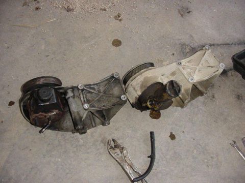 1992 Ford Bronco - Two serpentine-belt driven pumps (likely power steering pump housings) laid side by side on a concrete garage floor, one darker/greasy and…