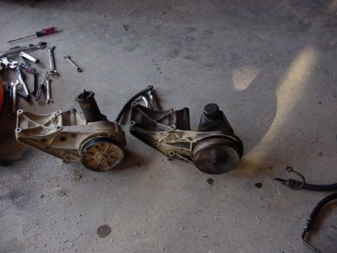 1992 Ford Bronco - Two automotive accessory brackets with pulleys (appear to be power steering pump assemblies) sit side by side on a concrete garage floor, w…