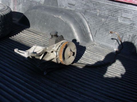 1992 Ford Bronco - A used power steering pump with a rusty pulley and attached metal and rubber lines, sitting in a black plastic truck bedliner with a dust s…