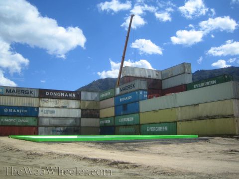 1990 Ford Bronco - Multicolored shipping containers stacked behind a green-edged concrete slab, with a crane boom against a bright blue sky.