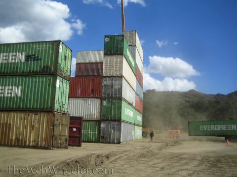 1990 Ford Bronco - Tall stacks of green, red and white shipping containers in a dusty dirt yard with a forklift and hazy hills behind.
