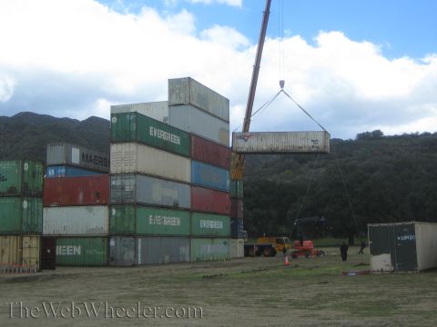 1990 Ford Bronco - mobile crane lowers a shipping container onto stacks of Evergreen and Maersk containers in a dirt yard ringed by hills.