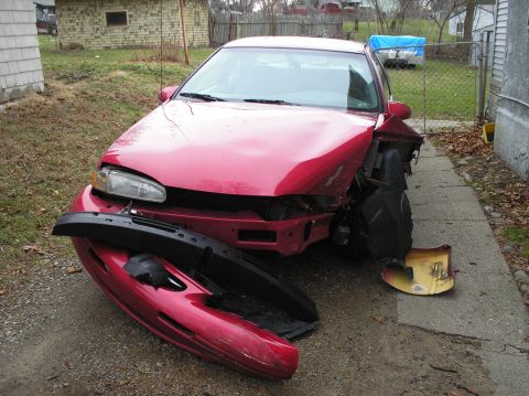 1995 Ford Thunderbird - A red sedan sits in a driveway with significant damage to its front end and a detached bumper.