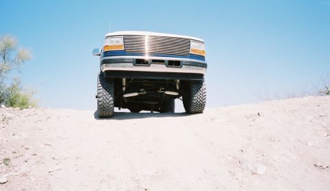 1996 Ford Bronco - A lifted Ford Bronco with a custom grille drives on a dirt hill under a clear blue sky.