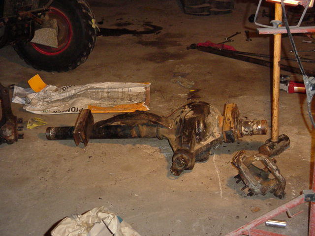 Removed solid front axle assembly lying on garage floor with leaf springs and steering knuckles