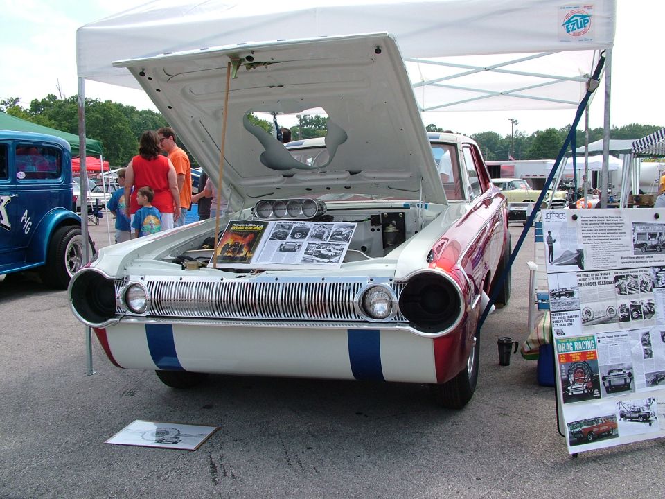 Front view of a white and red drag car with blue stripes, hood open showing engine and display boards next to it