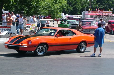 1972 Ford Ranchero - A bright orange muscle car with black racing stripes is parked at an outdoor event with other vehicles and spectators.