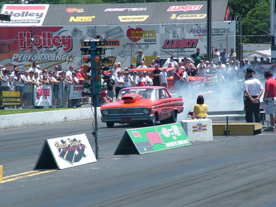 Red 1963 Ford Falcon drag car with a large hood scoop, emitting smoke