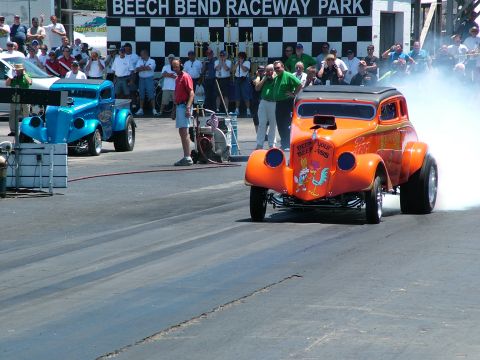 1972 Ford Ranchero - An orange hot rod car with cartoon graphics on the hood races down a track, kicking up smoke.