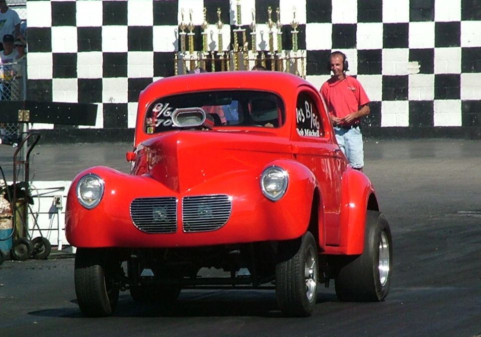 Red hot rod coupe with a scoop on the hood, parked at a drag strip with trophies in the background
