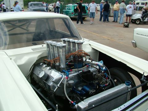 1972 Ford Ranchero - A highly modified V8 engine with six carburetors is visible under the hood of a white car, with other vehicles and people in the background.