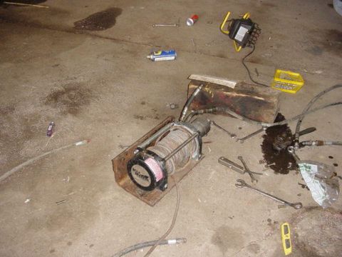 1992 Ford Bronco - A hydraulic winch with a steel-cable-wound drum sits removed on a garage floor, bolted to a rusty steel mounting plate with hydraulic hoses…