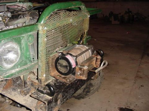 1992 Ford Bronco - A winch is mounted to the front bumper of a green vehicle, with a protective mesh guard.