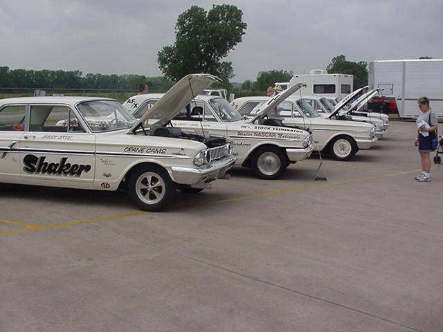 Several white vintage race cars lined up with hoods open. One car has 'Shaker' on the door