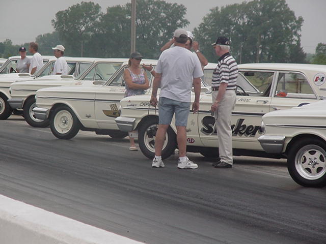 Several white vintage race cars lined up on a track, people standing around them